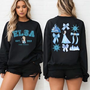 以下が含まれることがあります： 青い文字で「ELSA」と書かれた黒いスウェットシャツ。下に「EST. 2013」と「DISNEY PRINCESS」と書かれています。スウェットシャツの背面には、エルサ、リボン、雪の結晶、お城が青色で描かれています。