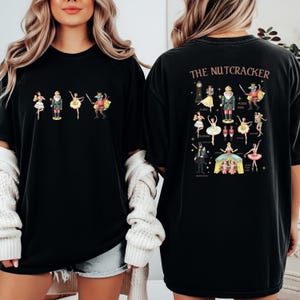 May include: Black t-shirt featuring a colorful illustration of characters from The Nutcracker ballet. The front of the shirt displays a row of characters, while the back showcases a detailed diagram with character names and a clock.