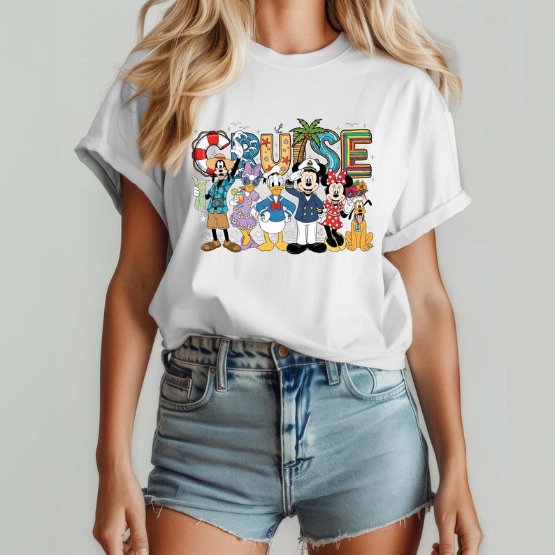 May include: White t-shirt featuring the word "CRUISE" in colorful, block letters with cartoon characters. The characters include Mickey Mouse, Minnie Mouse, Donald Duck, and Goofy. The shirt is paired with denim shorts.