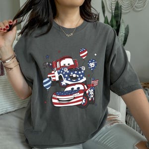 May include: A charcoal gray t-shirt featuring a graphic of two cartoon race cars decorated with red, white, and blue American flag patterns. The design includes balloons, one marked with the number 95, and small star accents surrounding the vehicles.