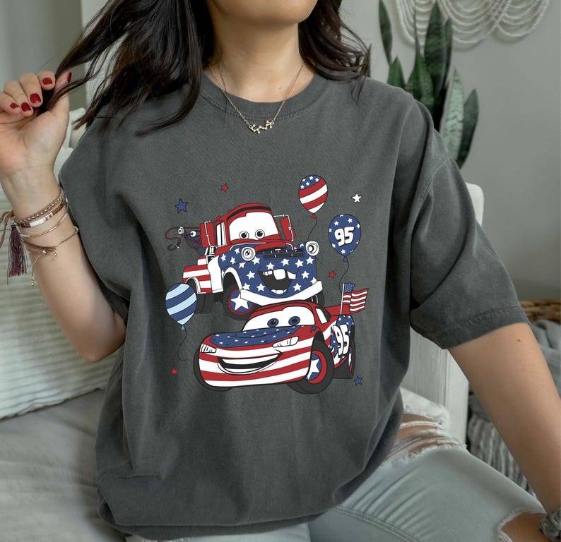 May include: A charcoal gray t-shirt featuring a graphic of two cartoon race cars decorated with red, white, and blue American flag patterns. The design includes balloons, one marked with the number 95, and small star accents surrounding the vehicles.