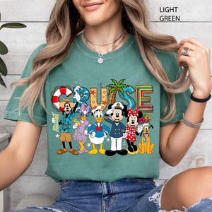 May include: Light green t-shirt featuring the word "CRUISE" in colorful letters with a palm tree and a life preserver. The shirt has a graphic of Disney characters including Mickey, Minnie, Donald, Daisy, Goofy, and Pluto.