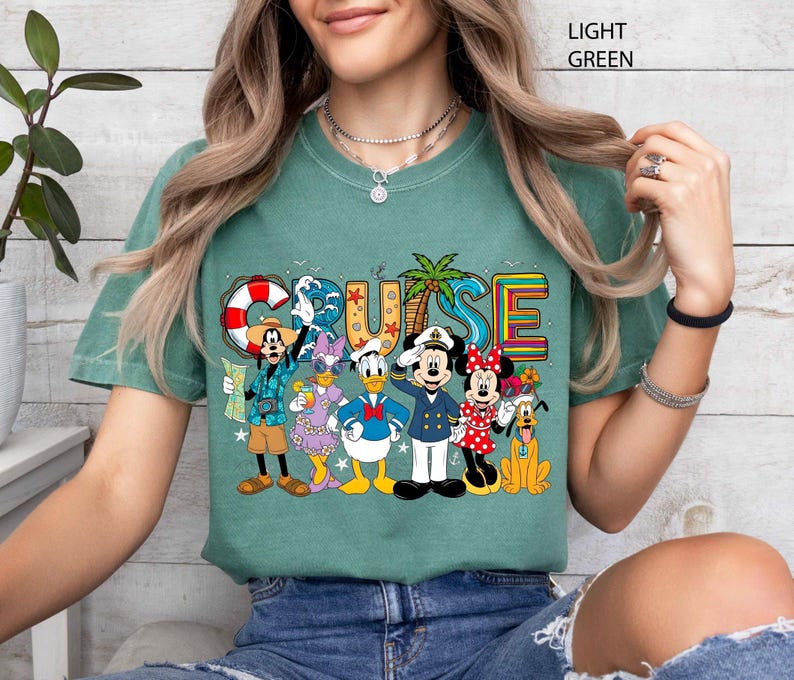 May include: Light green t-shirt featuring the word "CRUISE" in colorful letters with a palm tree and a life preserver. The shirt has a graphic of Disney characters including Mickey, Minnie, Donald, Daisy, Goofy, and Pluto.