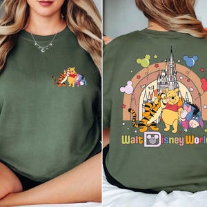 Op de afbeelding: Olijfgroene sweatshirt met een cartoonontwerp van Winnie de Poeh, Teigetje en Iejoor. De voorkant heeft een kleine afbeelding van de personages. De achterkant toont een regenboog, kasteel en de tekst "Walt Disney World."