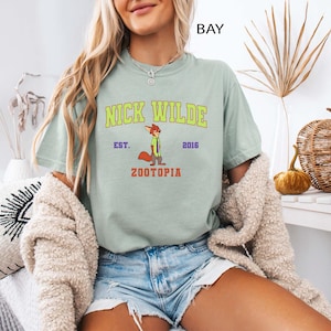 May include: A light green t-shirt featuring the text "NICK WILDE" in yellow and green, with a cartoon fox illustration. Below the text is "ZOOTOPIA" in red. The shirt is part of the Zootopia collection.