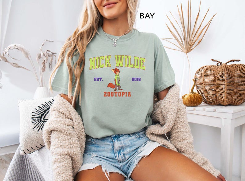 May include: A light green t-shirt featuring the text "NICK WILDE" in yellow and green, with a cartoon fox illustration. Below the text is "ZOOTOPIA" in red. The shirt is part of the Zootopia collection.