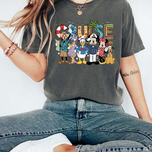 May include: Dark gray t-shirt with a colorful "CRUISE" graphic featuring Disney characters. The design includes Mickey Mouse, Minnie Mouse, Donald Duck, Goofy, and Pluto, with nautical elements like a life preserver and palm trees.