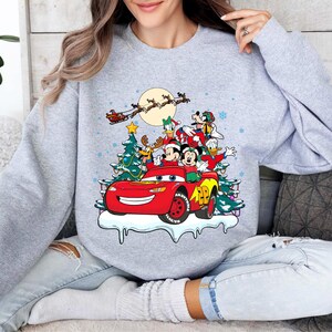 K&ouml;nnte beinhalten: Ein hellgraues Sweatshirt mit einem weihnachtlichen Design. Die Grafik zeigt ein rotes Rennauto mit Mickey und Minnie Mouse, Donald Duck und Goofy. Ein Weihnachtsbaum, Schneeflocken und der Schlitten des Weihnachtsmanns vervollst&auml;ndigen die Szene.