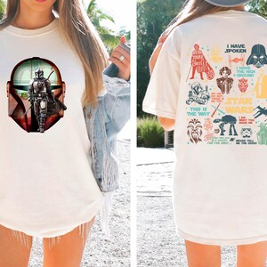 May include: Cream-colored t-shirt featuring a Mandalorian graphic on the front and Star Wars character illustrations on the back. The front graphic shows the Mandalorian in a helmet, with a green-skinned character. The back has various Star Wars icons and phrases.