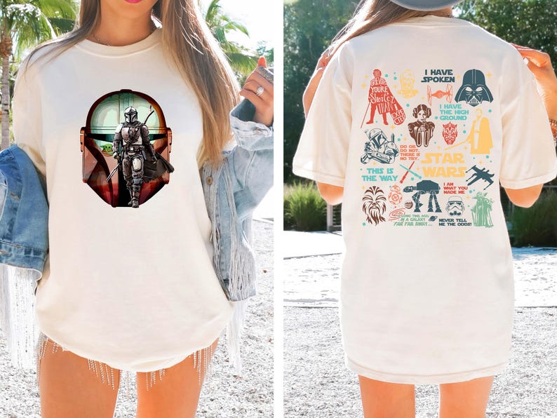May include: Cream-colored t-shirt featuring a Mandalorian graphic on the front and Star Wars character illustrations on the back. The front graphic shows the Mandalorian in a helmet, with a green-skinned character. The back has various Star Wars icons and phrases.