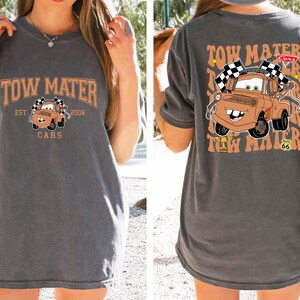 Puede incluir: Camiseta gris oscuro con una imagen de dibujos animados de Mate de la pel&iacute;cula Cars. La parte delantera de la camiseta tiene el texto "TOW MATER" encima de la imagen, con "EST. 2006" y "CARS" debajo. La parte trasera repite el texto y la imagen.