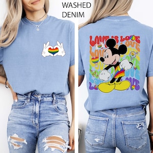 May include: Light blue t-shirt with a rainbow heart graphic made with white gloved hands. The back of the shirt features Mickey Mouse in rainbow colors with the words "Love is Love". The shirt is made of washed denim.