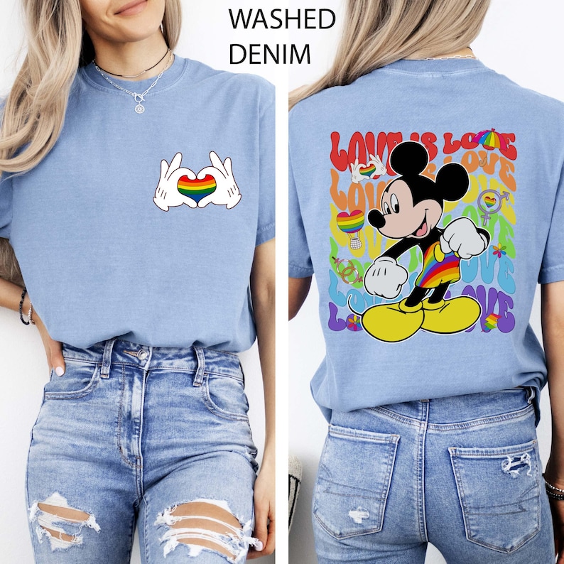 May include: Light blue t-shirt with a rainbow heart graphic made with white gloved hands. The back of the shirt features Mickey Mouse in rainbow colors with the words "Love is Love". The shirt is made of washed denim.