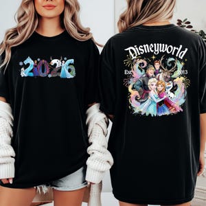 K&ouml;nnte beinhalten: Schwarzes T-Shirt mit dem Text "2026" in einem Schneeflocken-Design auf der Vorderseite. Auf der R&uuml;ckseite des Shirts steht "Disneyworld Est. 2013" mit einer farbenfrohen Grafik von Figuren aus dem Film Frozen.
