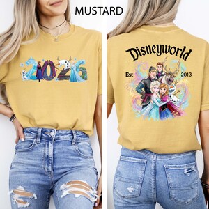 K&ouml;nnte beinhalten: Senffarbenes T-Shirt mit dem Text "2026" und Frozen-Charakteren auf der Vorderseite. Auf der R&uuml;ckseite steht "Disneyworld Est 2013" mit einer Grafik von Charakteren aus dem Film Frozen.