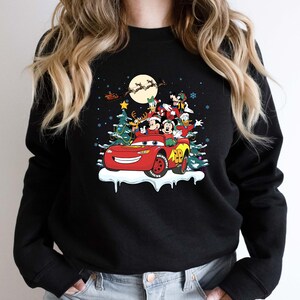 K&ouml;nnte beinhalten: Schwarzes Sweatshirt mit Rundhalsausschnitt und festlichem Aufdruck. Das Design zeigt ein rotes Auto mit Mickey Mouse, Donald Duck und anderen Figuren, dekoriert mit einem Weihnachtsbaum und Schnee. Der Weihnachtsmann und Rentiere sind im Hintergrund zu sehen, was ein Feiertags-Thema erzeugt.