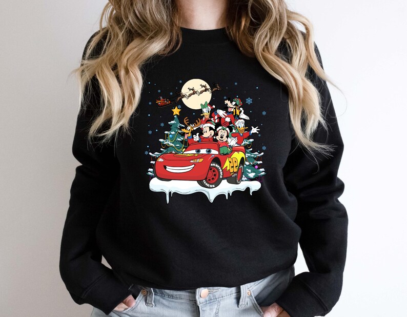 K&ouml;nnte beinhalten: Schwarzes Sweatshirt mit Rundhalsausschnitt und festlichem Aufdruck. Das Design zeigt ein rotes Auto mit Mickey Mouse, Donald Duck und anderen Figuren, dekoriert mit einem Weihnachtsbaum und Schnee. Der Weihnachtsmann und Rentiere sind im Hintergrund zu sehen, was ein Feiertags-Thema erzeugt.