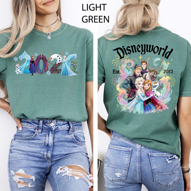 K&ouml;nnte beinhalten: Hellgr&uuml;nes T-Shirt mit Figuren aus dem Film Frozen. Auf der Vorderseite ist das Jahr "2026" mit den Figuren zu sehen. Auf der R&uuml;ckseite des Shirts steht "Disneyworld Est 2013" mit einer Grafik der Figuren.