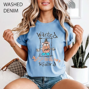 May include: Pale blue t-shirt featuring a cartoon potato character with the word "Wanted" above and "$50 Billion Reward" below. The illustration is framed. The words "Washed Denim" are in the top left corner. The shirt is a casual style.