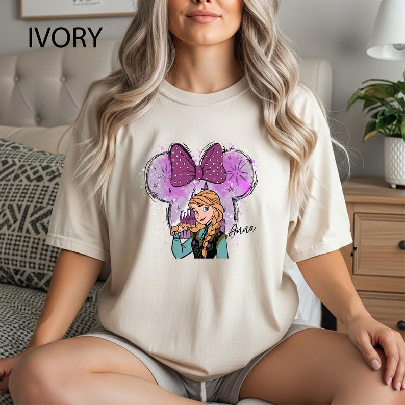 Pu&ograve; includere: T-shirt color avorio con una grafica di Anna di Frozen all'interno di un contorno di orecchie di Topolino. Il design include un fiocco viola, stelle e il nome "Anna". Un capo casual.