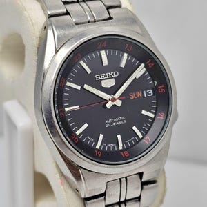 May include: A stainless steel Seiko 5 automatic wristwatch with a black dial. The watch features white hour markers, red accents, and a day-date display. The watch band is also stainless steel.
