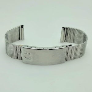 May include: A silver stainless steel watch strap with a mesh design. The strap has a rectangular clasp with the Omega logo and adjustment holes. The strap is designed to attach to a watch case.