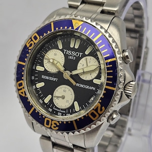 Peut inclure: Une montre chronographe Tissot en acier inoxydable avec un cadran noir, une lunette bleue et dorée, et un bracelet argenté. Le cadran affiche plusieurs sous-cadrans et le texte "TISSOT 1853". Résistant à l'eau jusqu'à 100 mètres.