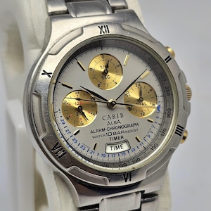 May include: A silver Carib Alba alarm-chronograph watch with a silver face and gold subdials. The watch has a stainless steel strap and a timer function. The watch face displays the text "CARIB ALBA ALARM-CHRONOGRAPH WATER 10 BAR RESIST TIMER TIME".