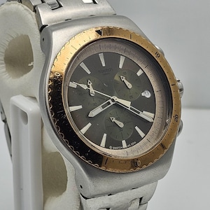 May include: A Swatch chronograph watch with a silver-tone case and band. The watch face is dark gray with gold-tone accents, including the bezel. The watch has three sub-dials and a date display at the 3 o'clock position. The text "Swatch" is visible.