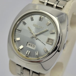 May include: A silver-toned Orient AAA wristwatch with a rectangular face. The watch features a light gray dial with silver hour markers, hands, and the day and date displayed. The watchband is silver and the watch is displayed on a white surface.