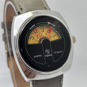 May include: A vintage Yema digital watch with a silver-tone case and a gray leather strap. The watch face is black with a yellow and green display showing hours and minutes. The watch reads "DIGITAL YEMA 17 Jewels".