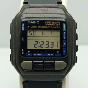 Op de afbeelding: Een vintage Casio Wrist Remote Controller horloge met een digitale display die de tijd en datum weergeeft. Het horloge is donkergroen met zwarte knoppen en een zwarte band. Op de wijzerplaat staat "CASIO WRIST REMOTE CONTROLLER ALARM CHRONOGRAPH".