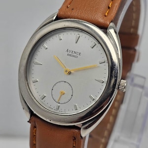 Peut inclure: Une montre-bracelet Avenue Seiko argentée avec un cadran clair, des aiguilles dorées et un petit cadran secondaire. La montre a un bracelet en cuir marron. Le cadran affiche le texte "AVENUE SEIKO".