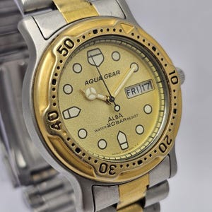 Puede incluir: Un reloj de pulsera Alba Aqua Gear dorado y plateado. La esfera del reloj es dorada con números negros y marcadores blancos. El reloj muestra el día y la fecha. La correa del reloj es plateada con detalles dorados. Resistente al agua hasta 20 bar.