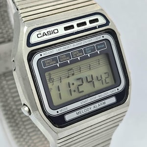 May include: A silver Casio digital watch with a rectangular face. The watch displays the time, date, and alarm functions. The watch face has the text "CASIO" and "MELODY ALARM". The watch band is made of silver metal.