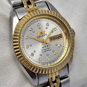 May include: A silver and gold-toned Orient Crystal watch with a silver dial. The watch features a gold-toned bezel and hands, with small crystal accents. The day and date are displayed in a window at the 3 o'clock position. The watch band is a combination of silver and gold links.