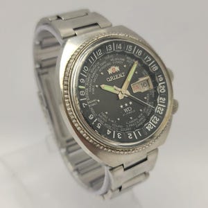 Puede incluir: Reloj de pulsera Orient WD 21 Jewels plateado con esfera negra. El reloj tiene un bisel de hora mundial, visualización de día y fecha y agujas luminosas. La correa del reloj también es plateada.