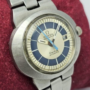 May include: A vintage Omega Genève Dynamic-C automatic wristwatch. The watch features a silver-tone stainless steel case and band. The watch face has a cream-colored center with a blue and white outer ring. The watch hands are blue, and the date is displayed at the 3 o'clock position.