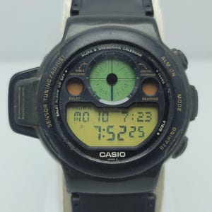May include: Black Casio digital watch with a green compass display and a yellow digital screen showing the time and date. The watch has multiple buttons and text including "HIJRA & GREGORIAN CALENDAR" and "CASIO JAPAN O".