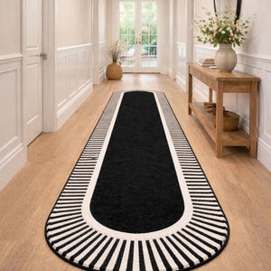Peut inclure: Tapis ovale noir et blanc rayé. Le centre est noir, bordé d'une bande blanche et de rayures noires et blanches. Le tapis est posé dans un couloir avec un sol en bois clair. Dimensions: environ 200 cm de long.