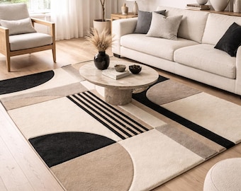 May include: A modern area rug with a geometric design in beige, black, and tan. The rug features a mix of shapes, including circles, rectangles, and stripes. The rug is placed in a living room with a sofa, armchair, and coffee table.