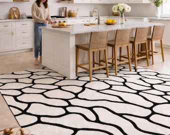 Puede incluir: Una gran alfombra de color crema con un llamativo patrón orgánico negro domina el primer plano de una cocina luminosa. El diseño de la alfombra presenta formas irregulares interconectadas. La cocina tiene gabinetes blancos, una encimera de mármol y taburetes de bar tejidos.