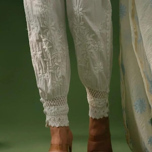 Puede incluir: Pantalones blancos bordados con motivos florales y ribete de encaje en los tobillos. Los pantalones forman parte de un conjunto y se combinan con tacones de tiras transparentes. La tela parece ligera.