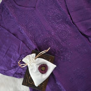 May include: A deep purple long-sleeved tunic featuring intricate tonal floral embroidery around the neckline and front. A small white drawstring pouch with a dark purple flower accent sits atop a dark wooden block placed on the fabric.