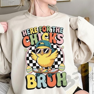 Könnte beinhalten: Beiges Sweatshirt mit farbenfrohem Aufdruck. Der Aufdruck zeigt "HERE FOR THE CHICKS BRUH" und zeigt ein gelbes Küken mit Sonnenbrille und blauer Kappe auf einem Skateboard, mit Ostereiern und einem karierten Hintergrund.