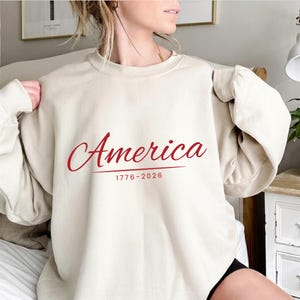 May include: A beige crewneck sweatshirt featuring the word America in a red cursive script font, with the text 1776-2026 printed in a smaller red sans-serif font directly underneath the word, centered on the front of the garment.