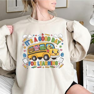 May include: A beige crewneck sweatshirt featuring a graphic of a yellow school bus with a bear, rabbit, frog, and duck inside. The text reads IT'S A GOOD DAY FOR LEARNING, surrounded by colourful illustrations of pencils, clouds, and stars.