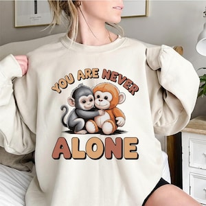 Puede incluir: Sudadera beige con un gráfico de dos monos de dibujos animados abrazándose. El texto "YOU ARE NEVER ALONE" está encima de los monos, con "ALONE" debajo. El diseño utiliza tonos marrones y naranjas.