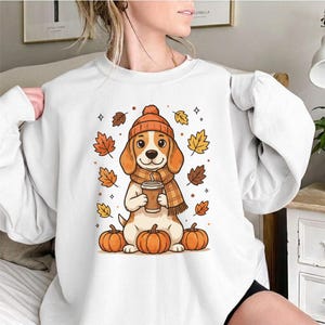 May include: A white crewneck sweatshirt featuring a graphic illustration of a beagle wearing an orange knit hat and a matching plaid scarf. The dog holds a coffee cup, surrounded by falling autumn leaves and three orange pumpkins at the base.