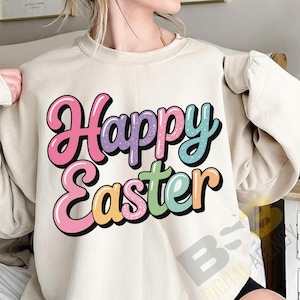 May include: A cream-coloured sweatshirt featuring the text "Happy Easter" in a colourful, bubble-style font. The text uses pink, purple, blue, green, and orange shades, with a black outline. The sweatshirt has long sleeves.